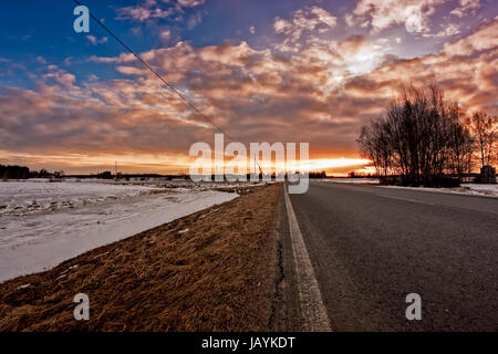 Einer ruhigen Straße im ländlichen Finnland. Die Frühlingssonne setzt schön hinter den Bäumen und Farben am Himmel auf dramatische Weise. Stockfoto