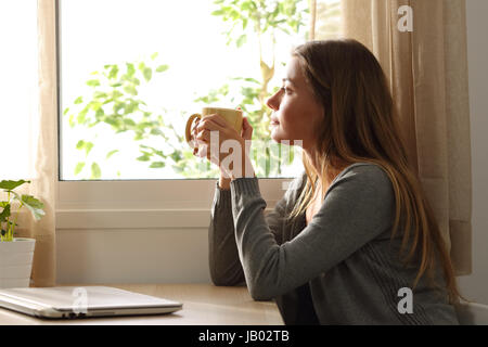 Seitenansicht einer entspannten Frau Blick durch ein Fenster in einem Tisch sitzen und eine Tasse Kaffee zu Hause zu halten Stockfoto