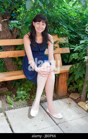 eine junge Frau sitzt auf einer Bank in einem Garten durch einen canicular Sonnentag Stockfoto