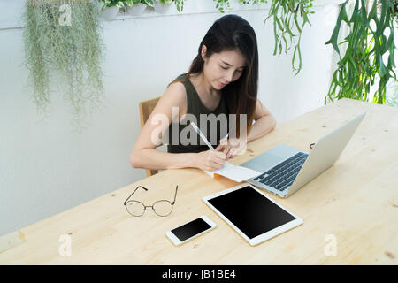 Bürotisch mit jungen asiatischen Frau schreiben auf Papier Notizblock, Asiatin freiberuflich zu Hause am Arbeitsplatz, Konzept der jungen Frau mit Laptop. Stockfoto