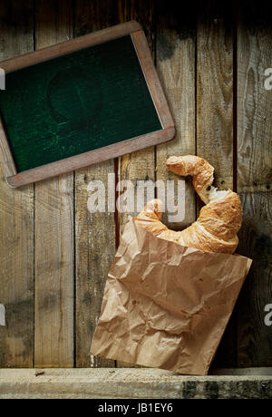 Draufsicht auf eine frisch gebackene golden Croissant, eine Tasse voll Röstkaffee Espresso und alte leere Schule Schiefer für ein Menü oder einen Text auf urigen Holzbrettern Stockfoto