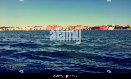 Linie der Gebäude auf der gegenüberliegenden Insel gegen das klare blaue Meer und den Himmel in Venedig, Italien Stockfoto