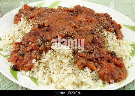 Nahaufnahme von vegetarisches Essen Bio Oregano Kraut, gewürfelte Tomaten, Kidney-Bohnen, schwarze Bohnen und Pinto über brauner Reis auf einem runden weißen Teller mit lackierten Bambus-Blätter auf ein grünes Tischset gelegt. Stockfoto