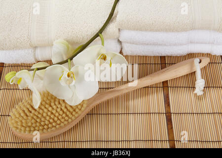 Ein Bad Pinsel liegt auf einer Matte aus Bambus. Im Hintergrund sind zwei Haufen gefaltete Handtücher mit eine weiße Orchidee geschmückt. Stockfoto
