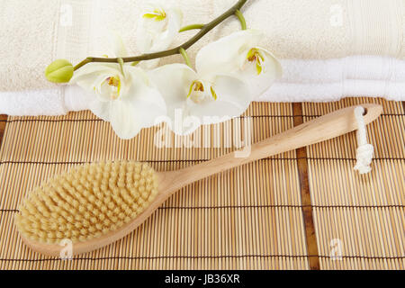 Ein Bad Pinsel liegt auf einer Matte aus Bambus. Im Hintergrund sind zwei Haufen gefaltete Handtücher mit eine weiße Orchidee geschmückt. Stockfoto