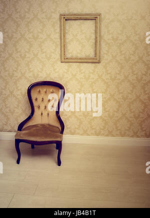Antik Polsterstuhl in einem tapezierten Zimmer mit eine leere Vintage Holz Bilderrahmen hängt an der Wand geeignet als ein Innenverzieren-Hintergrund Stockfoto