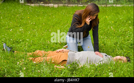 Schöne Frau, die erste Hilfe für ein hübscher junger Mann und verwenden ihr Handy den Krankenwagen, Reanimation, auf einer Wiese rufen zurück Stockfoto