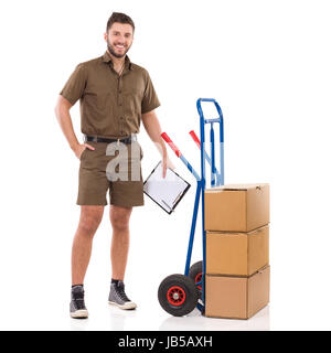 Männliche Kurier steht in der Nähe ein Push Cart holding Zwischenablage und Blick in die Kamera. Voller Länge Studio gedreht isoliert auf weiss. Stockfoto