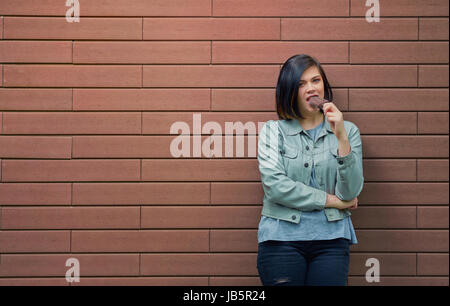 Junge schöne Mädchen Schokolade Eis am Stiel Essen. Frau auf einer gemauerten Wand Hintergrund auf der Straße Stockfoto