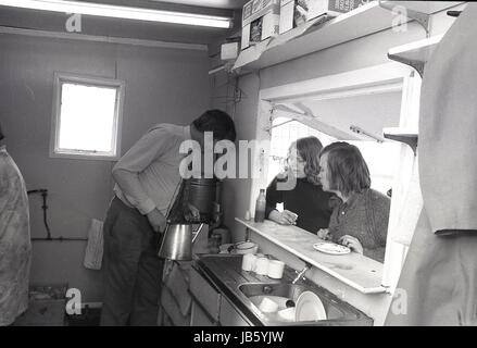 4 Pence für eine Tasse Tee im berühmten Blackheath Tea Hut, Greenwich, London, SE3 1973, Historisches Bild von zwei Kunden, die draußen unter dem Baldachin an der Theke des Blackheath Tea Hut mit ihren Tassen Tee standen, Während der männliche Server den Teekocher aus einer metallenen Wasserkanne füllt. Die einfache Snackbar mit Servierluke war ein bekannter Raststopp für Reisende auf der A2 Straße in Süd-London. Stockfoto