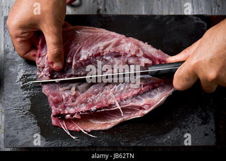 High-Angle Shot eines kaukasischen Jünglings schneiden eine rohe frische Makrele mit einem Messer auf einem Schiefer Tablett platziert auf einem rustikalen Holztisch oder Arbeitsplatte Stockfoto