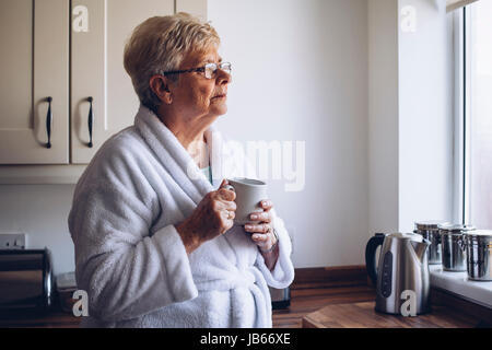 Ältere Frau aus ihrem Küchenfenster schauen mit einer Tasse Tee in der hand. Stockfoto