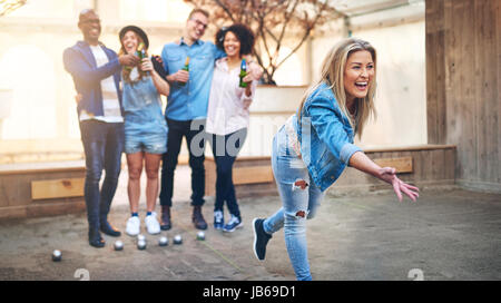 Junge begeistert Frau wirft einen Ball für Petanque während schwarze und weiße Freunde klirrende Flaschen Bier und Spaß haben. Stockfoto
