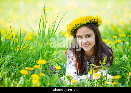 Hübsche junge Mädchen mit Kranz von Löwenzahn und lächelnd, während auf Gras im Park Stockfoto