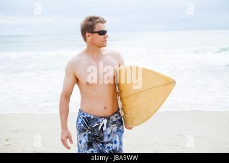 Entspannt und konzentriert aussehende männliche Surfer zu Fuß am Strand während des Tragens Surfbrett Stockfoto