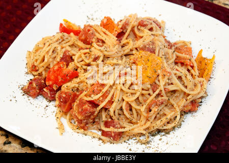 Gesunden bereit Hartweizen Weizen Grieß-Spaghetti mit gehackten roten und orangefarbenen Paprikaschoten gemischt, gewürfelte Tomaten, geriebener Parmesan, Knoblauch und gekrönt mit trocken Basilikum Kraut auf einem quadratischen weißen Teller. Stockfoto