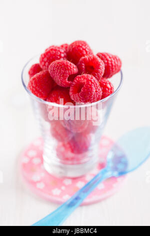 Nahaufnahme von frischen roten Himbeeren im Glas mit blauen Plastiklöffel ruht auf kleinen rosa Teller auf der Leuchtfläche Stockfoto