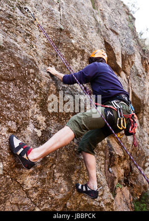 Ein junger Mann mit Helm und Seil klettern sicher eine Felswand im Freien. Stockfoto