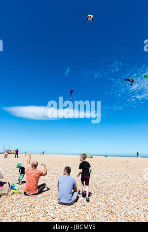 Menschen auf stoney Strand in Walmer, Deal, in Kent, England sitzen während Drachen fliegen. Verschiedene Drachen fliegen im blauen Himmel. Stockfoto