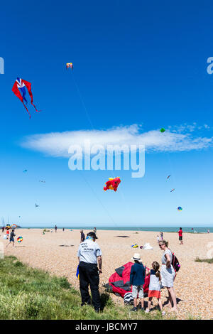 Menschen auf stoney Strand in Walmer, Deal, in Kent, England sitzen während Drachen fliegen. Verschiedene Drachen fliegen im blauen Himmel. Stockfoto