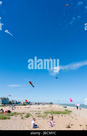 Menschen auf stoney Strand in Walmer, Deal, in Kent, England sitzen während Drachen fliegen. Verschiedene Drachen fliegen im blauen Himmel. Stockfoto