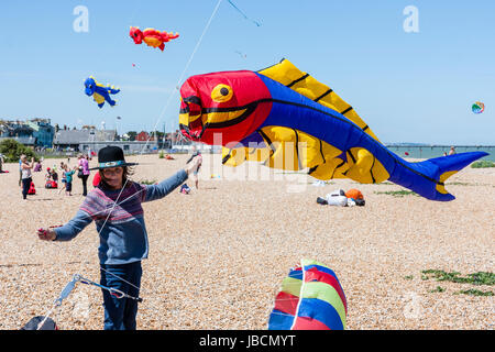 Mann auf stoney Strand in Walmer, in Kent, England, hält einen großen Fisch geformt Kite über es zu starten. Andere Kites in verschiedenen Formen im Hintergrund fliegen. Drachen fliegen. Stockfoto