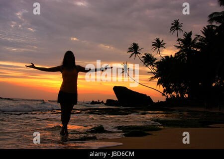 Silhouette Frau an einem Strand mit Palmen und Felsen bei Sonnenuntergang, Unawatuna, Sri Lanka Stockfoto