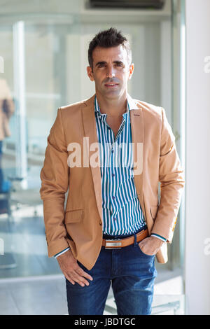 Ein 35-40 Jahre alte Mann kaukasischen dunkle Haare cool modernes Aussehen mit Hand in der Tasche in eine abgespeckte Shirt, Blazer und Jeans im Büro, mittlere totale Stockfoto