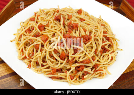 Sehr lecker aussehende Abendessen perfekt gekochte Spaghetti Nudeln Nudeln gemischt mit Bio Tomatenwürfel und Kräuter, die auf einem weißen Teller mit schönen Intarsien aus Holz Serviertablett auf einer roten Tischdecke serviert. Stockfoto