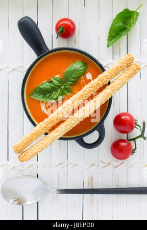 Draufsicht auf eine Tomatensuppe in einer schwarzen Topf, beträufelt mit Sahne und garniert mit frischem grünen Basilikum und lange Brot-Sticks, auf weißem Hintergrund aus Holz Stockfoto