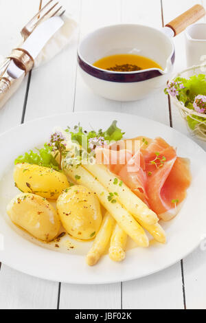 Gekochten Spargel mit italienischen Schinken und gekochte Babykartoffeln serviert mit Olivenöl und Kräuter dressing auf einer weißen hölzernen Theke in einer Landhausküche Stockfoto