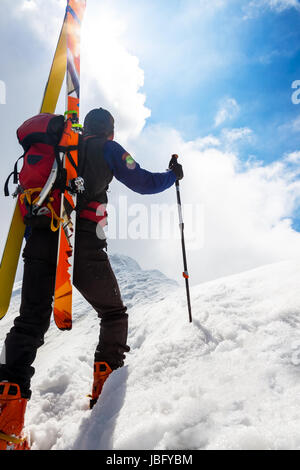 Skibergsteiger entlang einer steilen verschneiten Grat mit den Skiern in den Rucksack. Im Hintergrund ein dramatischer Himmel mit einem glänzenden hellen Sonne. Konzepte: Abenteuer, Erfolg, Mut, Entschlossenheit, Selbstverwirklichung, gefährliche Tätigkeit, Extremsport, Winter Freizeit. Stockfoto