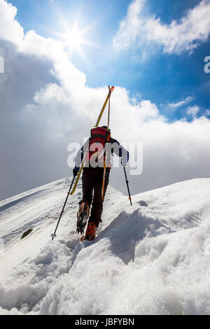 Skibergsteiger entlang einer steilen verschneiten Grat mit den Skiern in den Rucksack. Im Hintergrund ein dramatischer Himmel mit einem glänzenden hellen Sonne. Konzepte: Abenteuer, Erfolg, Mut, Entschlossenheit, Selbstverwirklichung, gefährliche Tätigkeit, Extremsport, Winter Freizeit. Stockfoto