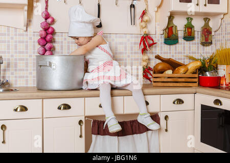 Kleines Mädchen mit Schürze und Kappe der Koch in der Küche im Haus sitzen. Mutters Helfer. 2 Jahre alt. Stockfoto