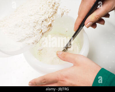 Weibliche Person Mehl in die geschmolzene Mischung aus Butter und Eigelb mit einem schwarzen Spatel mischen Stockfoto