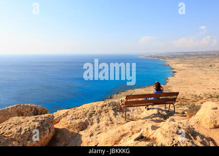 Eine einsame Frau sitzen auf einer Bank am Kap Greco Klippe Top Blick über das Mittelmeer in Richtung Ayia Napa in der Ferne. Zypern. Stockfoto