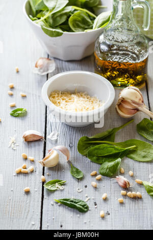 Produkte für die Herstellung von selbstgemachtem Pesto: Blattspinat, geriebener Käse, Knoblauch, Pinienkerne, Olivenöl und Blender auf weißem Holz Hintergrund Stockfoto
