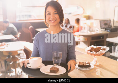 Porträt lächelnd Kellnerin mit Tablett mit Cappuccino, Brownie und Wasser im café Stockfoto