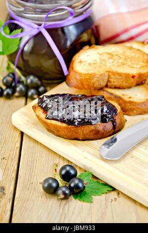 Scheiben geröstetes Brot, ein Glas mit schwarzen Johannisbeeren Marmelade, Messer auf Holzbrett Hintergrund Stockfoto