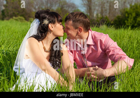 Porträt von Braut und Bräutigam küssen Lippen beim Sitzen auf der Wiese Stockfoto