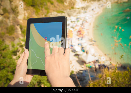Weibliche Hände halten digitale Tablet-Gerät mit GPS-Navigation-Map-Anwendung auf dem Hügel oberhalb des wunderschönen Strandes. Urlaub und Reisen-Konzepte. Stockfoto