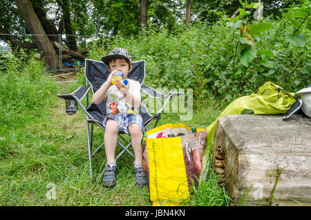 Ein junger Bot sitzt in einem Klappstuhl auf einem Campingplatz und Snjoys einen Drink. Stockfoto