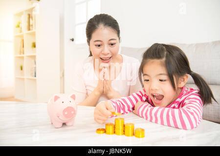wenig asiatische chinesisches Mädchen Kinder gespannt zu sehen, wie viel Geld sie Einzahlung in ihr Sparschwein mit ihrer Mutter in das Wohnzimmer hat zu Hause. Familie acti Stockfoto