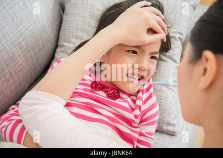 Junge Mutter Überprüfung Temperatur des Kranken wenig asiatische chinesische Tochter liegend auf dem Sofa im Wohnzimmer zu Hause. Familientätigkeit Konzept. Stockfoto