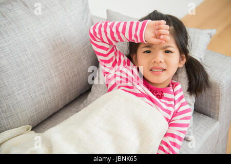 Kleine chinesische Asiatin mit Krankheit und Hand auf ihrem Kopf fühlen sich unwohl auf Sofa im Wohnzimmer zu Hause. Familientätigkeit Konzept. Stockfoto