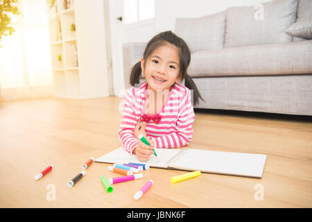 Glückliches Kind spielt. Kleines Kind Mädchen zieht mit Farbstiften liegend auf dem Holzboden im Wohnzimmer zu Hause. Familientätigkeit Konzept. Stockfoto