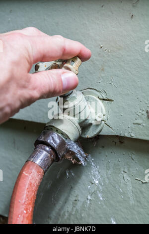 Vertikale Foto einer Reifen kaukasischen männliche Hand outdoor Wasserhahn Drehgriff mit Wasser aus, wo der Schlauch befestigt Stockfoto