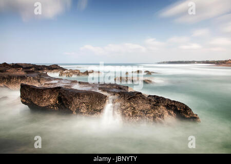 Lange Exposition Seelandschaft mit Mini-Wasserfälle, die auf einem Felsen im Meer an der balinesischen Küste mit beeindruckendem Wolken und herrliche Panorama-Aussicht Stockfoto