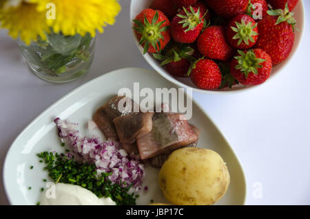 Eingelegter Hering mit neuen Kartoffeln, Schnittlauch, roten Zwiebeln und Sauerrahm auf einem Porzellanteller. Eine Schale mit Erdbeeren und Mittsommer Blumen auf der Seite. Stockfoto