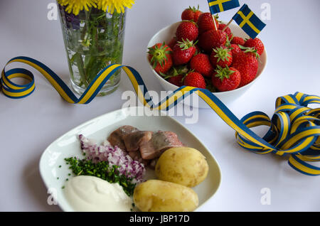 Eingelegter Hering mit neuen Kartoffeln, Schnittlauch, roten Zwiebeln und Sauerrahm auf einem Porzellanteller. Eine Schüssel Erdbeeren mit schwedischen Flaggen und Mittsommer flowe Stockfoto